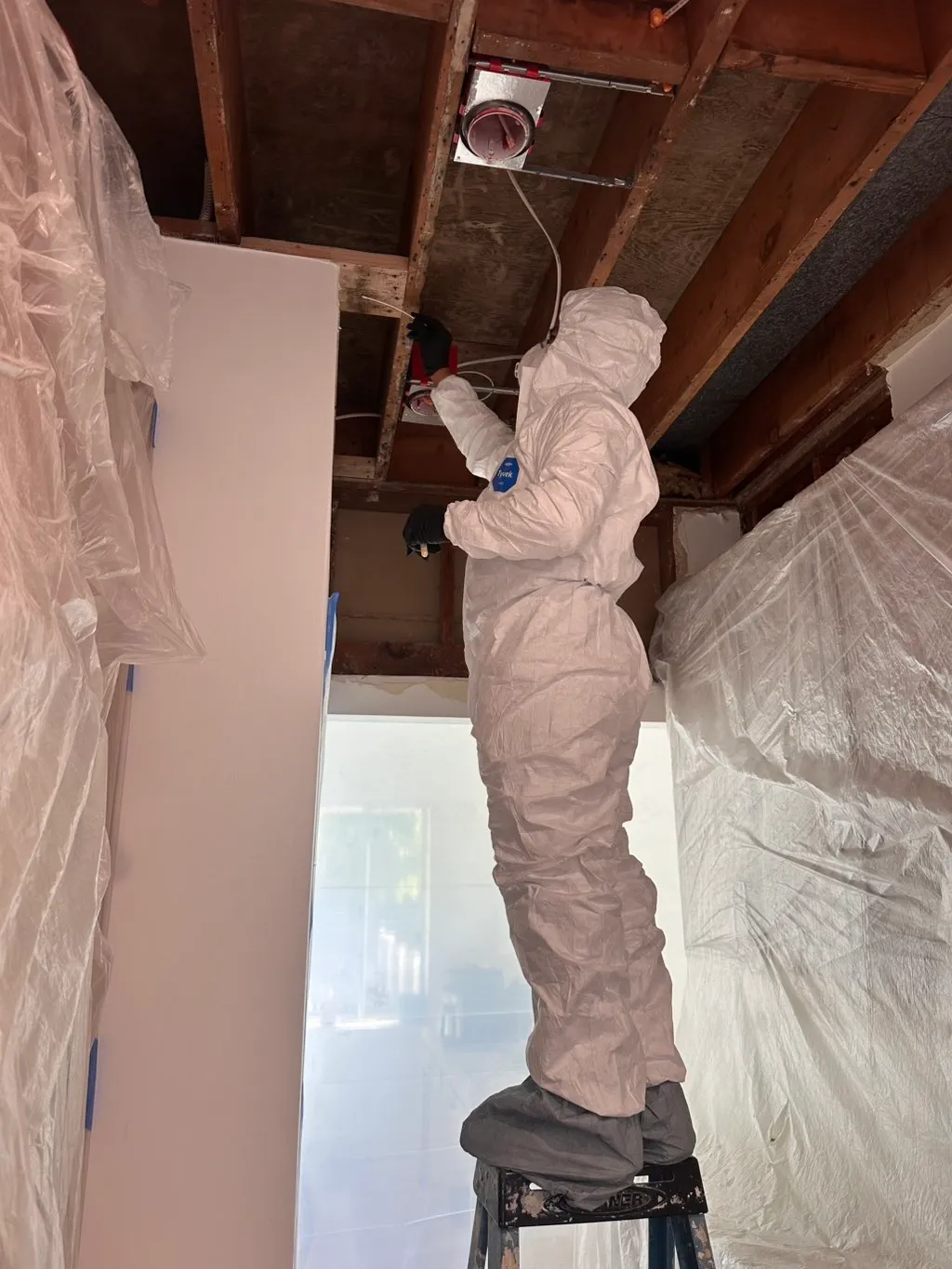 Certified technician in PPE performing ceiling demolition behind containment plastic.
